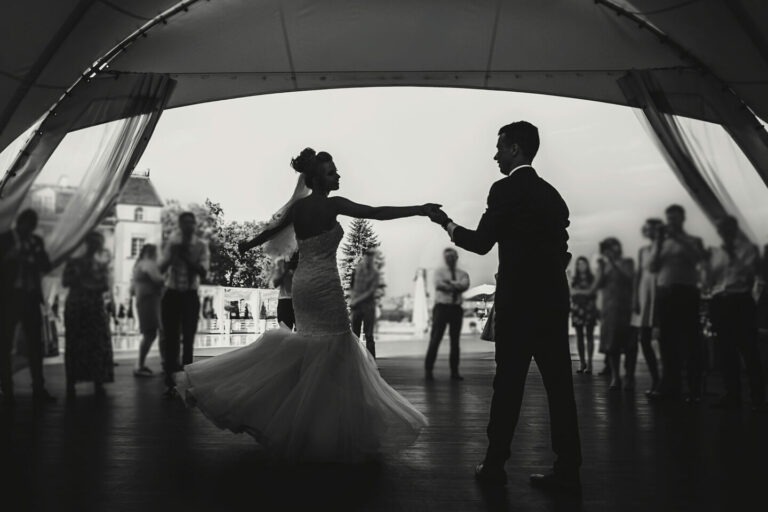 Silhouettes of happy bride and groom gently dancing at wedding reception. Gorgeous wedding couple of newlyweds embracing while having first dance on background of guests